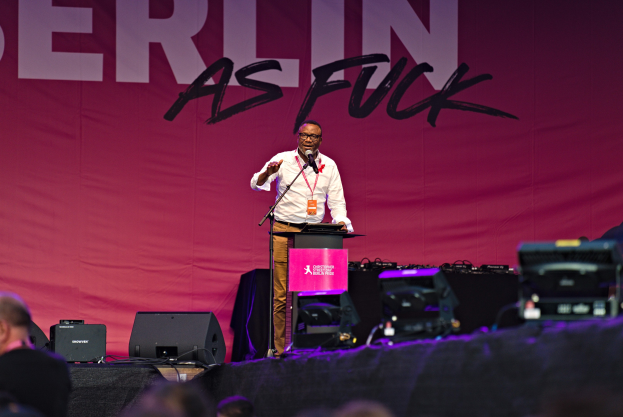 Mann in weißem Hemd und braunen Hosen, der in ein Mikrofon spricht, vor einem pinken "Berlin as Fuck"-Plakat auf der Bühne.