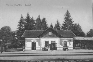 Ein altes Schwarz-Weiß-Foto eines Bahnhofs in Warmbad Villach, Bahnhof, mit einem Haus mit Fenstern und Türen, einer Gruppe von Menschen, die auf Bänken stehen und sitzen, einem Zaun, einem Straßenpfahl, einem Schild, einigen Bäumen und einem bewölktem Himmel.