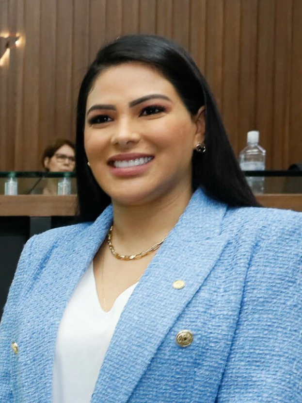 Eine Frau in einer blauen Blazer und weißem Hemd, identifiziert als Bürgermeisterin von San Antonio Ana Ortiz, steht lächelnd vor einer hölzernen Wand mit zwei Personen an einem Tisch dahinter, beleuchtet von einem Licht.