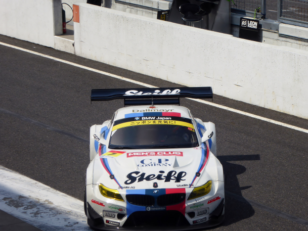 Ein BMW M6 GT3 Rennauto auf einer Rennstrecke mit Text auf dem Auto, einer Wand im Hintergrund und Maschendrahtzaun.