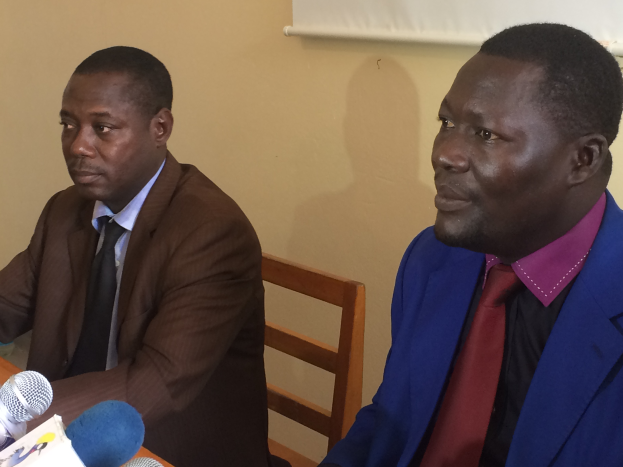 Zwei M├Ąnner in Blazern sitzen an einem Tisch mit Mikrofonen, Papieren und anderen Gegenst├Ąnden und f├╝hren ein Gespr├Ąch gegen eine cremefarbene Wand mit einem wei├čen Objekt.