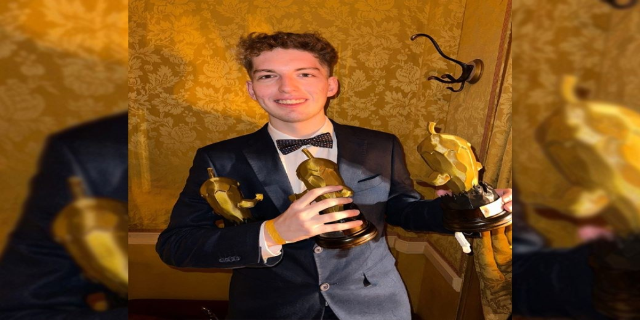 Ein Mann im Smoking hält eine Oscar-Statue und lächelt, mit einer Wand im Hintergrund und einem Wasserzeichen auf dem Bild.