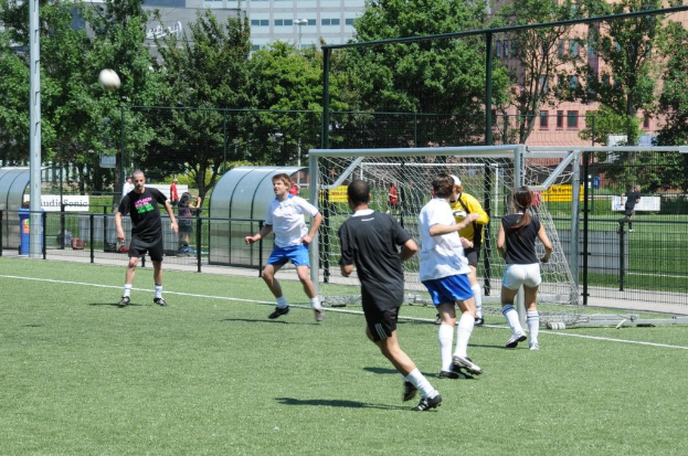 Personen in Sportbekleidung beim Fußballspielen auf einem Feld mit einem Zaun und einem Tor, einem Pfosten und einem Ball oben links und Bäumen und Gebäuden im Hintergrund.