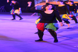 Eine Gruppe junger Frauen tanzt auf einer Bühne, im Hintergrund ist ein Gemälde an der Wand zu sehen, alle tragen Schuhe.