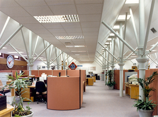 Leerer Büroraum mit Reihen von Schreibtischen und Stühlen, Topfpflanzen, Säulen, einer Wanduhr und ordentlich angeordneten Deckenleuchten.