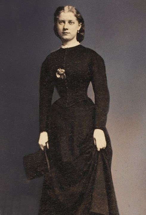 Ein Schwarz-Weiß-Foto einer älteren Frau in einem langen schwarzen Kleid mit hohem Kragen und langen Ärmeln, die mit ernster Miene, gefalteten Händen und einem ordentlichen Dutt steht und eine Tasche hält.