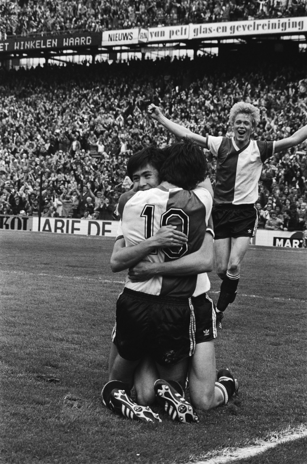 Drei Fußballspieler umarmen sich auf dem Boden nach einem Tor, mit jubelnder Menge und schwenkenden Fahnen im Hintergrund, in Schwarz-Weiß aufgenommen.