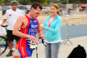 Ein Mann in einem roten und blauen Triathlonanzug läuft eine Straße entlang, neben ihm geht eine Frau, im Hintergrund sind Menschen, Fahrräder, Verkehrshütchen, Geländer, Fahrzeuge, Bäume und andere Gegenstände zu sehen.