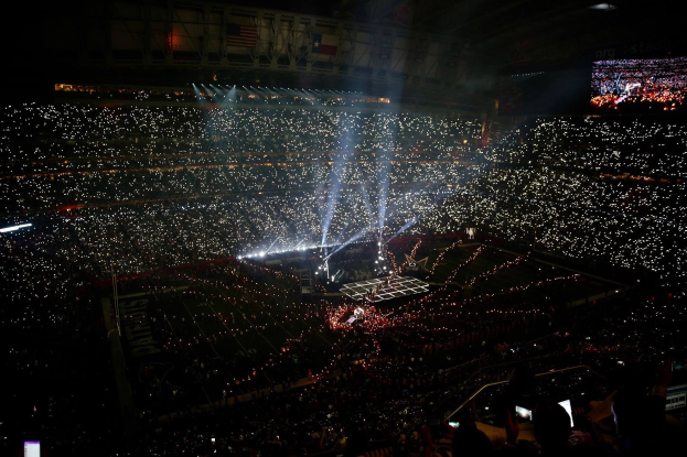 Eine große Menge in einem hell erleuchteten Stadion bei Nacht schaut auf einen Bildschirm auf der rechten Seite, der die Ergebnisse der Super Bowl XLVII-Halbzeitshow anzeigt.