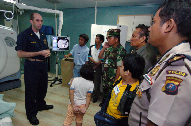 Gruppe von Menschen in einem Krankenhauszimmer mit einem Mann, der ein Mikrofon hält, einem drapierten Bett, medizinischer Ausrüstung, einem Röntgengerät, einer Texttafel, einer Tür und Deckenleuchten.