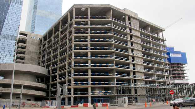 Großes Bauprojekt in einer Stadt mit Skyscrapern im Hintergrund, mit Fahrzeugen, Fußgängern, Verkehrsinfrastruktur und Kränen unter einem klaren Himmel.