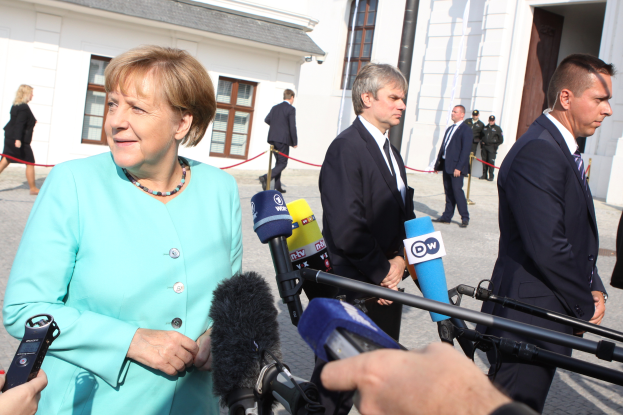 Bundeskanzlerin Angela Merkel spricht vor der Kanzleramt in Berlin mit Reportern, umgeben von Menschen mit Mikrofonen, mit Gebäuden im Hintergrund.