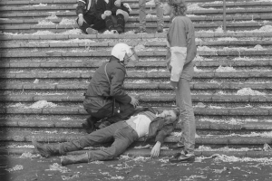 Eine Schwarz-Weiß-Szene eines Mannes, der neben einem Polizeibeamten auf dem Boden liegt, mit einigen Menschen im Hintergrund, die stehen und auf einer Treppe sitzen.