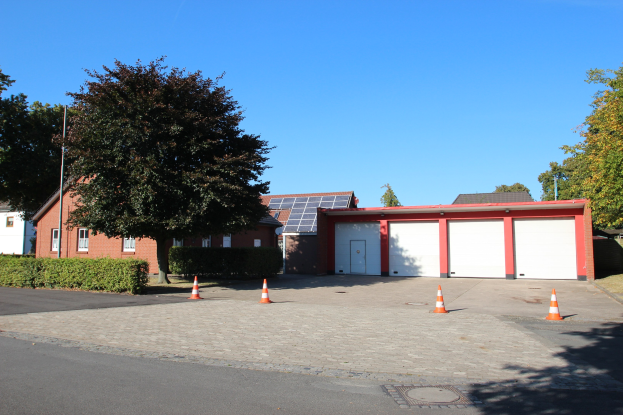 Straße mit Verkehrskegeln, die zu einem Gebäude mit Solarpanelen auf dem Dach führt, umgeben von Bäumen und Pflanzen unter einem klaren blauen Himmel.