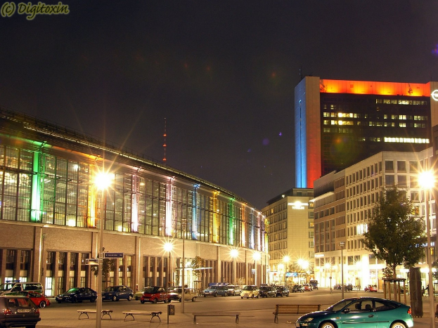 Eine nächtliche Stadtansicht mit beleuchteten Gebäuden, Laternen, Bäumen, Bänken und Fahrzeugen auf der Straße, mit Text in der oberen linken Ecke.