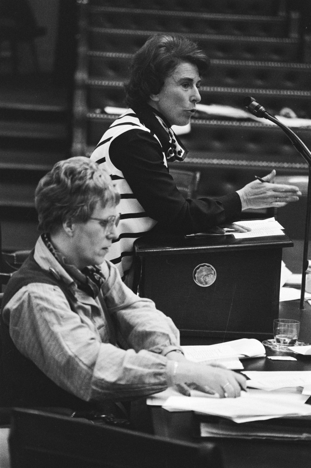 Schwarz-weiß-Foto einer Frau, die an einem Tisch im Europäischen Parlament in Straßburg im Mai 1981 sitzt und einen Stift hält, mit Papieren, einem Glas Wasser und anderen Gegenständen auf dem Tisch, mit einer Treppe im Hintergrund.