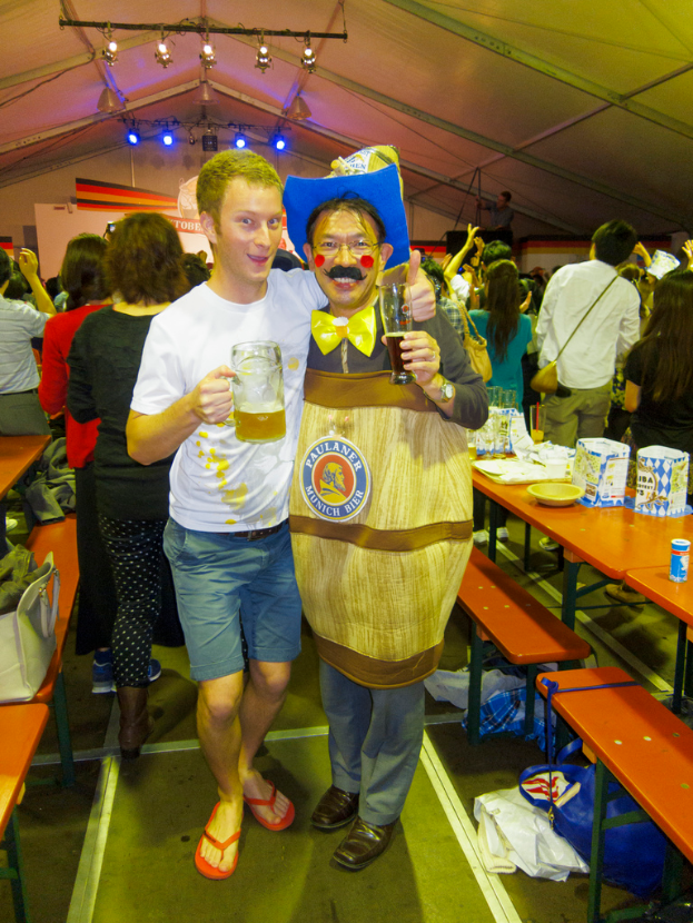 Zwei Personen stehen zusammen, eine hält ein Kind mit einem Bier, die andere trägt ein Dramakostüm, mit einem Tisch voller Essen in der Nähe und einer Gruppe von Menschen dahinter.