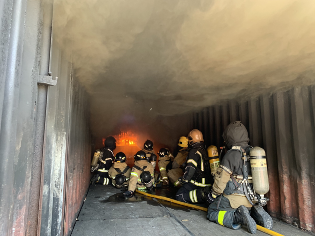 Feuerwehrleute in Schutzausrüstung, einschließlich Helmen und Sauerstoffflaschen, arbeiten in einem raucherfüllten Tunnel mit einem sichtbaren Feuer im Hintergrund, einer hält ein Rohr und eine Wand ist links zu sehen.