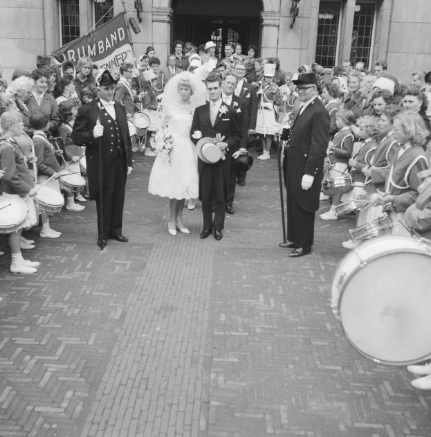 Eine Braut in einem weißen Kleid und Schleier und ein Bräutigam mit einem Strauß gehen eine Straße entlang vor einer Menge, darunter einige mit Musikinstrumenten, mit einem Gebäude und einem Banner im Hintergrund, dargestellt in Schwarz-Weiß.