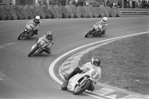 Eine Gruppe von Menschen in Helmen fährt Motorräder auf einer Rennstrecke, die von Gras, Pflanzen und einem Maschendrahtzaun umgeben ist, mit Zuschauern und Texttafeln im Hintergrund.