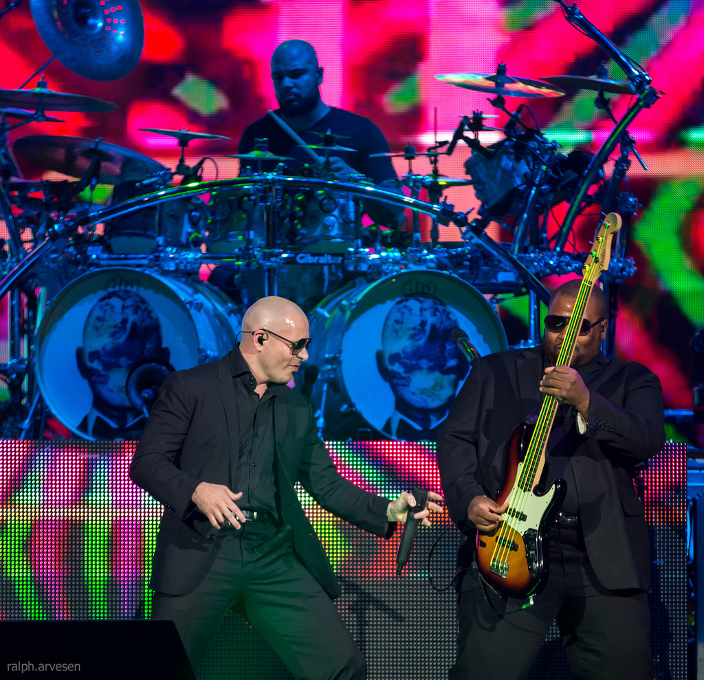 Ein Mann auf der rechten Seite spielt eine Gitarre, ein anderer Mann in einem schwarzen Blazer hält ein Mikrofon und lächelt, während ein Mann in einem schwarzen T-Shirt Schlagzeug spielt, alles vor einem bunten Hintergrund bei einem Musikkonzert.