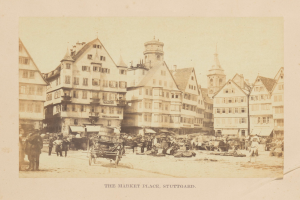 Altes Schwarz-Weiß-Foto eines belebten Marktplatzes in Stuttgart, Deutschland, mit mehrstöckigen Gebäuden, Passanten, horse-drawn carts und verschiedenen Marktständen; handschriftlicher Text erscheint entlang der unteren Kante.