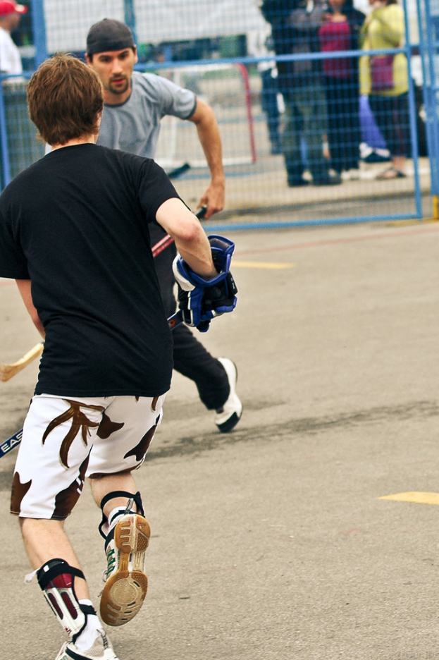 Zwei Menschen spielen Hockey auf einer Straße mit einem Zaun dahinter und einer Menge von Menschen im Hintergrund.