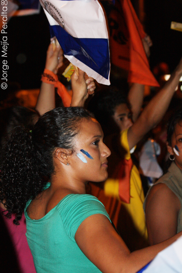 Eine Frau in einem gr├╝nen T-Shirt steht auf einer Demonstration und h├Ąlt ein wei├čes Schild in der Hand, hinter ihr stehen viele andere Menschen.