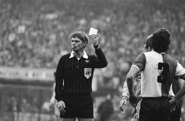 Ein Schiedsrichter in Schwarz und Weiß hält eine rote Karte vor einer Menge von Menschen in Fußballtrikots hoch, mit einem Zaun im Hintergrund.