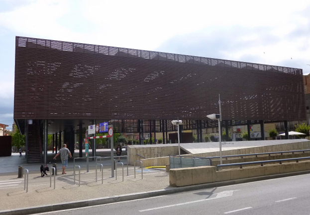 Großes modernes Gebäude mit zahlreichen Fenstern als neuer städtischer Eingang, umgeben von städtischer Infrastruktur einschließlich Straßenlaternen, Fahrzeugen, Fußgängern und Bäumen unter einem bewölkten Himmel.