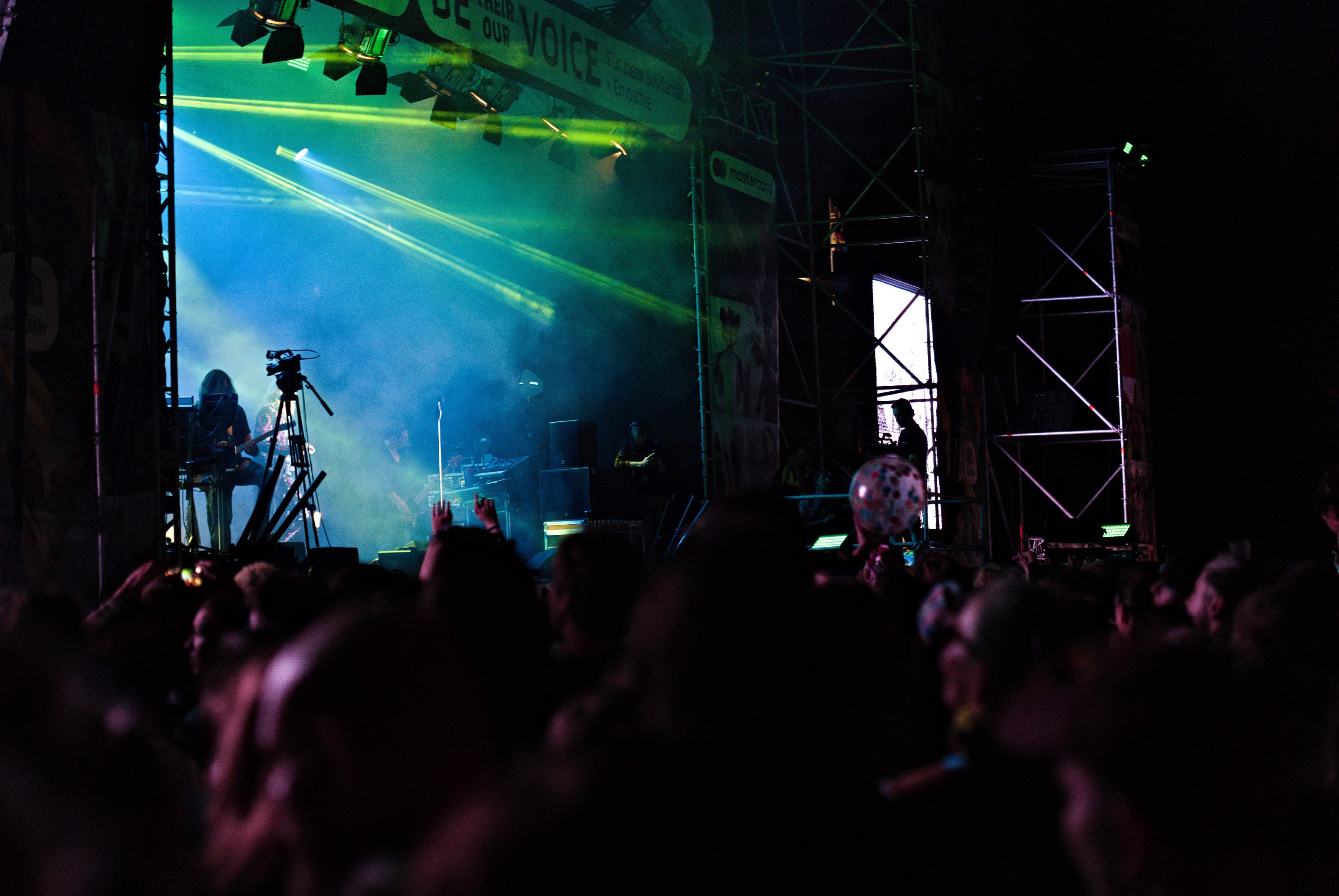 Eine Menge Menschen auf einem Konzert schaut auf eine hell erleuchtete Bühne mit Musikinstrumenten, Mikrofonen und einem Banner im Hintergrund.