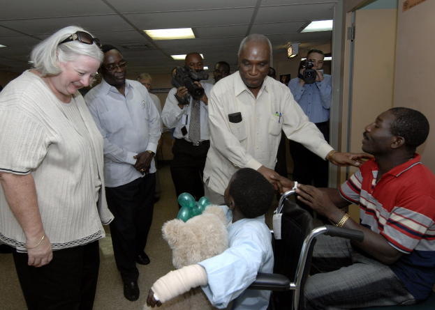 Eine Gruppe von Menschen mit Kameras umringt ein Kind im Rollstuhl, das ein weißes Hemd trägt und einen Verband am Arm hat, in einem Raum mit Deckenbeleuchtung und einer Tür im Hintergrund.