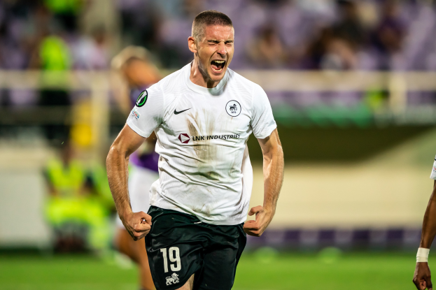 Ein Mann in weißem Hemd und schwarzen Shorts läuft auf einem Fußballfeld, feiert ein Tor, umgeben von einer Gruppe von Menschen, mit einem unscharfen Hintergrund.