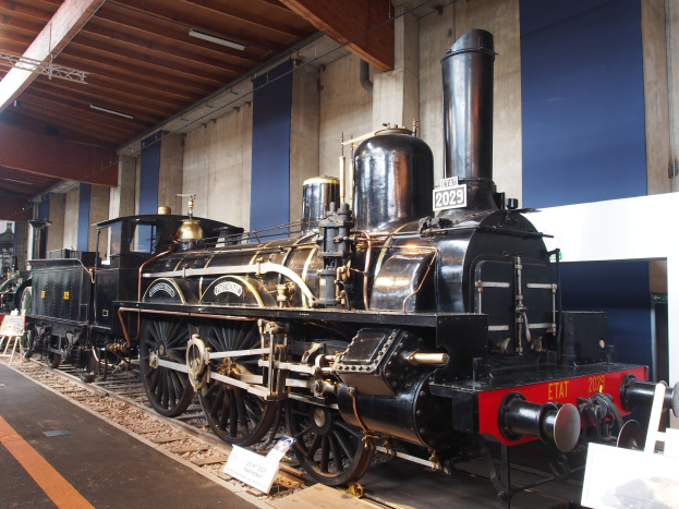 Alte Dampflokomotive in einem Museums Setting mit umliegenden Eisenbahnschienen, Informationsschildern und anderen Ausstellungsstücken, vor einer Wand mit Deckenbeleuchtung.