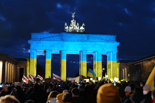 Eine Menschenmenge steht vor dem Brandenburger Tor in Berlin, hält Fahnen und Schilder, mit einerBanner auf der rechten Seite.