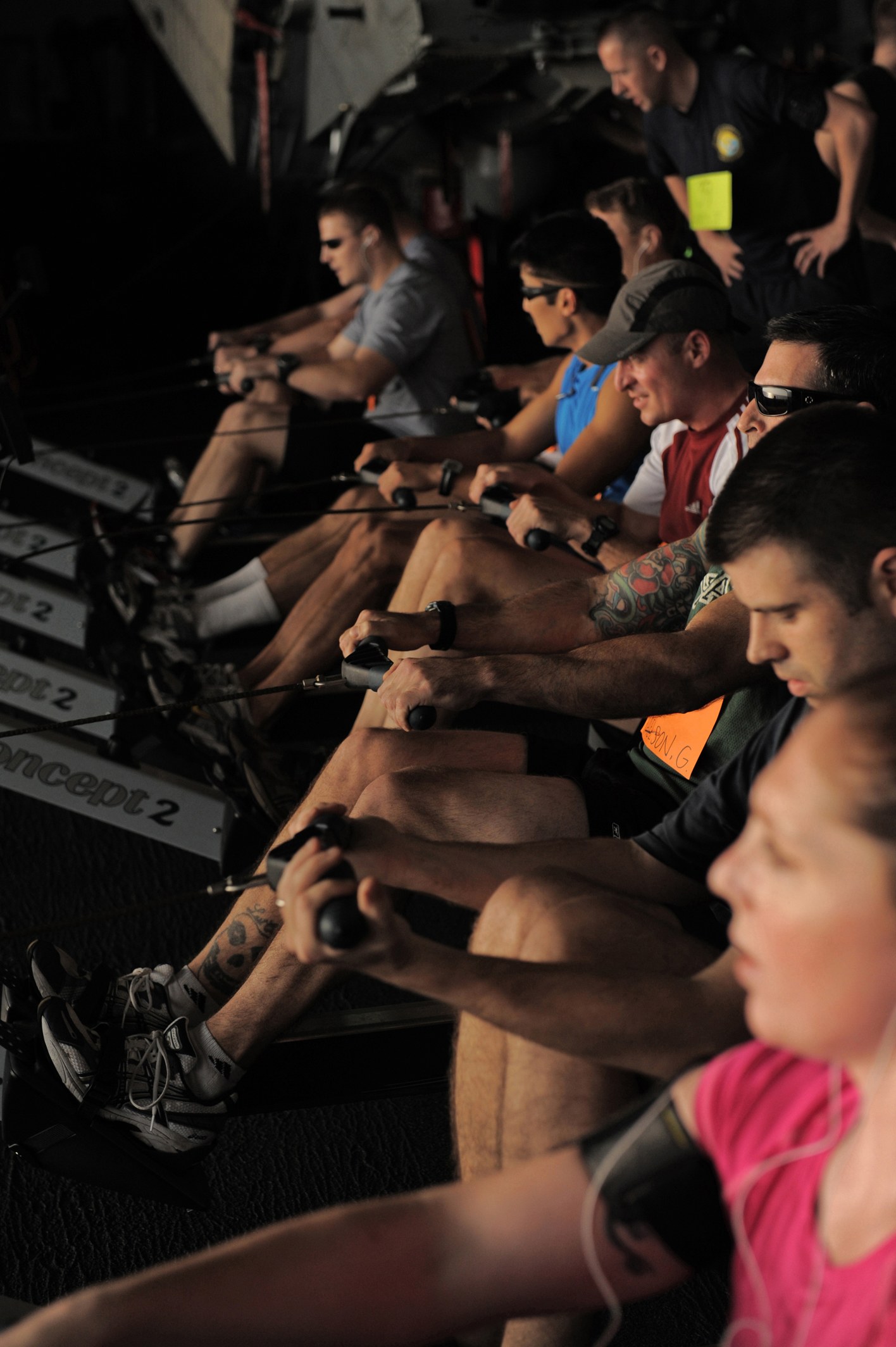 Gruppe von Menschen, die auf Rudermaschinen in einem Fitnessstudio sitzen, mit einigen im Hintergrund, die alle Sportschuhe tragen.