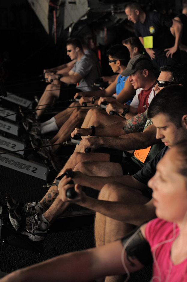 Gruppe von Menschen, die auf Rudermaschinen in einem Fitnessstudio sitzen, mit einigen im Hintergrund, die alle Sportschuhe tragen.