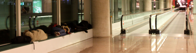 Menschen auf einer Bank in einem Flughafen mit zwei geraden Rolltreppen im Hintergrund.