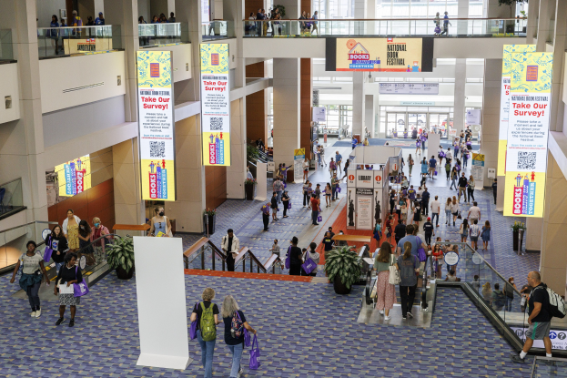 Eine große Gruppe von Menschen geht durch ein großes Gebäude mit Schildern, Topfpflanzen, Rolltreppen, Geländern, Säulen, bedruckten Tafeln, Glastüren und anderen Gegenständen, einige tragen Taschen, wahrscheinlich nehmen sie am Nationalen Buchfest teil.