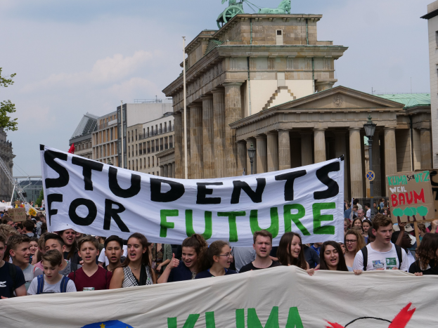 Gruppe von Schülern marschiert in Berlin mit einem bunt bemalten 'Schüler für die Zukunft' Banner vor Gebäuden, Bäumen und Himmel.