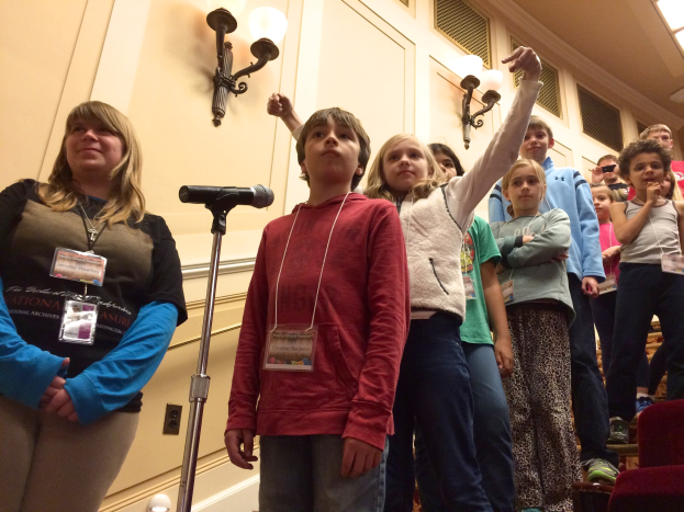 Gruppe von Kindern, die vor einem Mikrofon singen, mit einer Frau daneben, Stühlen auf der rechten Seite und Wandlampen im Hintergrund.