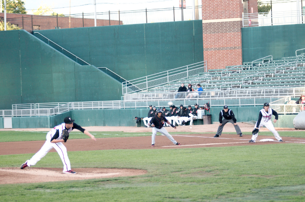 Baseball players are on a field playing a game, with spectators seated on benches and chairs behind a railing, cars, a wall, pillars, and trees in the background.