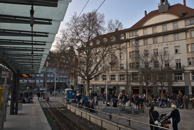 Eine belebte Straßenkreuzung mit einem Bahnhof auf der linken Seite, umgeben von Gebäuden, Bäumen, Laternenmasten und Texttafeln, wo Menschen gehen, auf Bänken sitzen oder stehen.