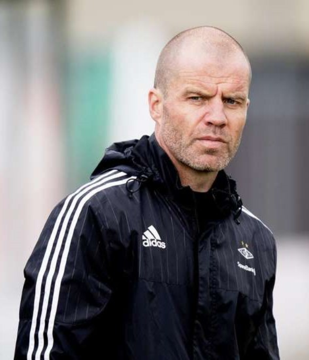 Ein kahlköpfiger Trainer in einer schwarzen Jacke steht auf einem Fussballfeld, trägt eine marineblaue Jacke mit weissen Streifen und einem weissen Kragen, die Hände sind verschränkt und der Gesichtsausdruck ernst.