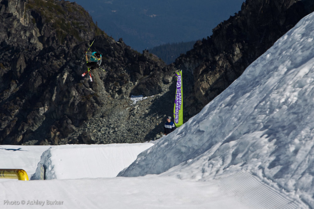 Ein Mann in Skier springt in der Luft mit schneebedeckten Bergen und einer Flagge mit Text im Hintergrund, und ein weiterer Mann steht vor der Flagge, mit Text in der unteren linken Ecke.
