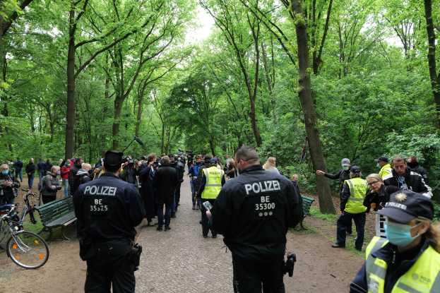 Polizisten vor einer Menschenmenge bei einer Outdoor-Veranstaltung mit Fahrrädern und einer Bank im Vordergrund und Bäumen im Hintergrund.