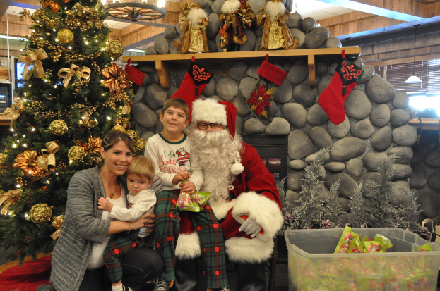 Eine Frau, Kinder und ein als Santa Claus verkleideter Mann mit Brille und Handschuhen stehen in der Nähe eines geschmückten Weihnachtsbaums, einer Wand mit Felsen, Spielzeug, Dekorationen und einem Korb mit Gegenständen, mit Lichtern im Hintergrund.