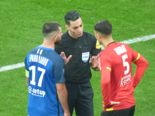 Ein Schiedsrichter in schwarzer Kleidung spricht mit zwei Männern auf einem Fussballfeld, einer in blauer und roter Shirts mit Text, beide tragen Brillen.