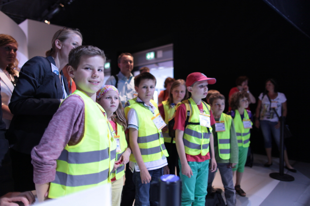 Eine Gruppe von Kindern trägt Sicherheitswesten und Namensschilder und steht auf einem Boden, wobei ein Junge links lächelt, ein Pfahl rechts steht und einige Menschen im Hintergrund zu sehen sind.