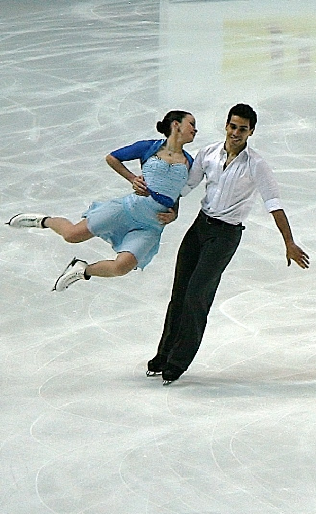 Ein Mann in einem weißen Hemd und schwarzen Hosen und eine Frau in einem blauen Kleid tanzen gemeinsam auf einem Eisstadion, ihre Arme und Beine ineinander verschlungen, in perfekter Synchronisation.